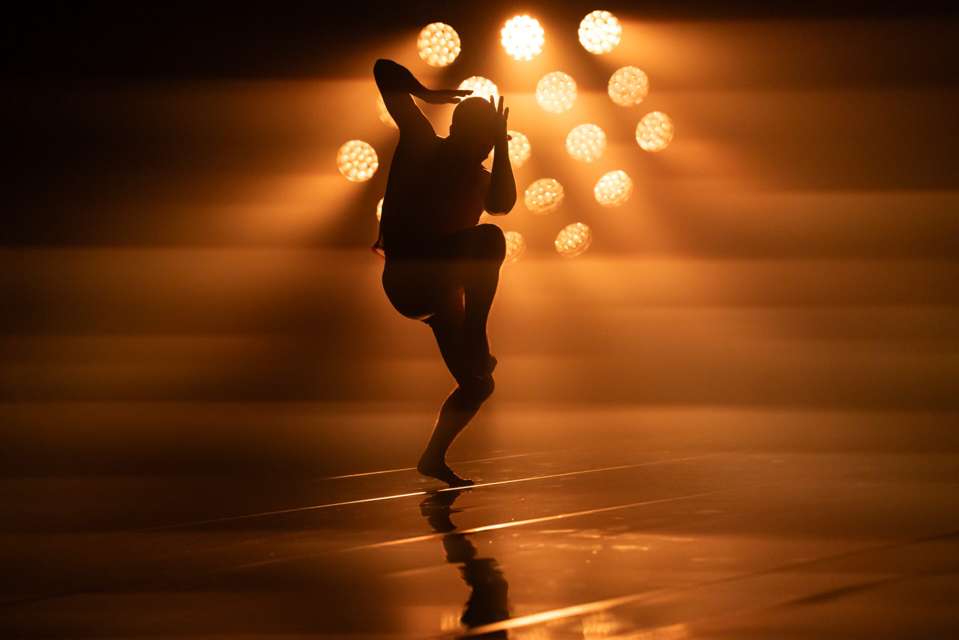 A single dancer bathed in orange light poses on a single leg with his arms close to his body.