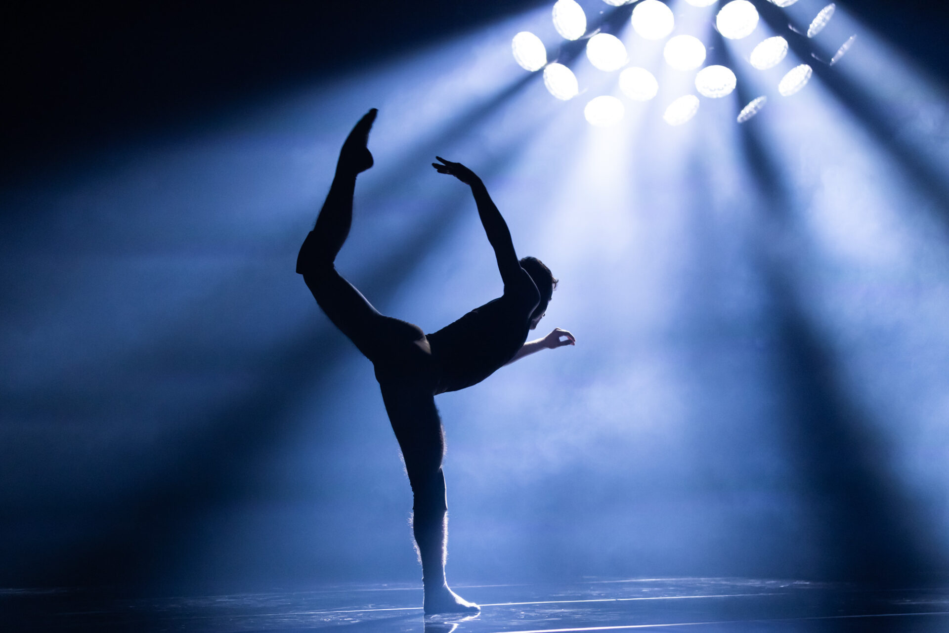 A single dancer bathed in pale blue light extends their foot behind them towards the ceiling.