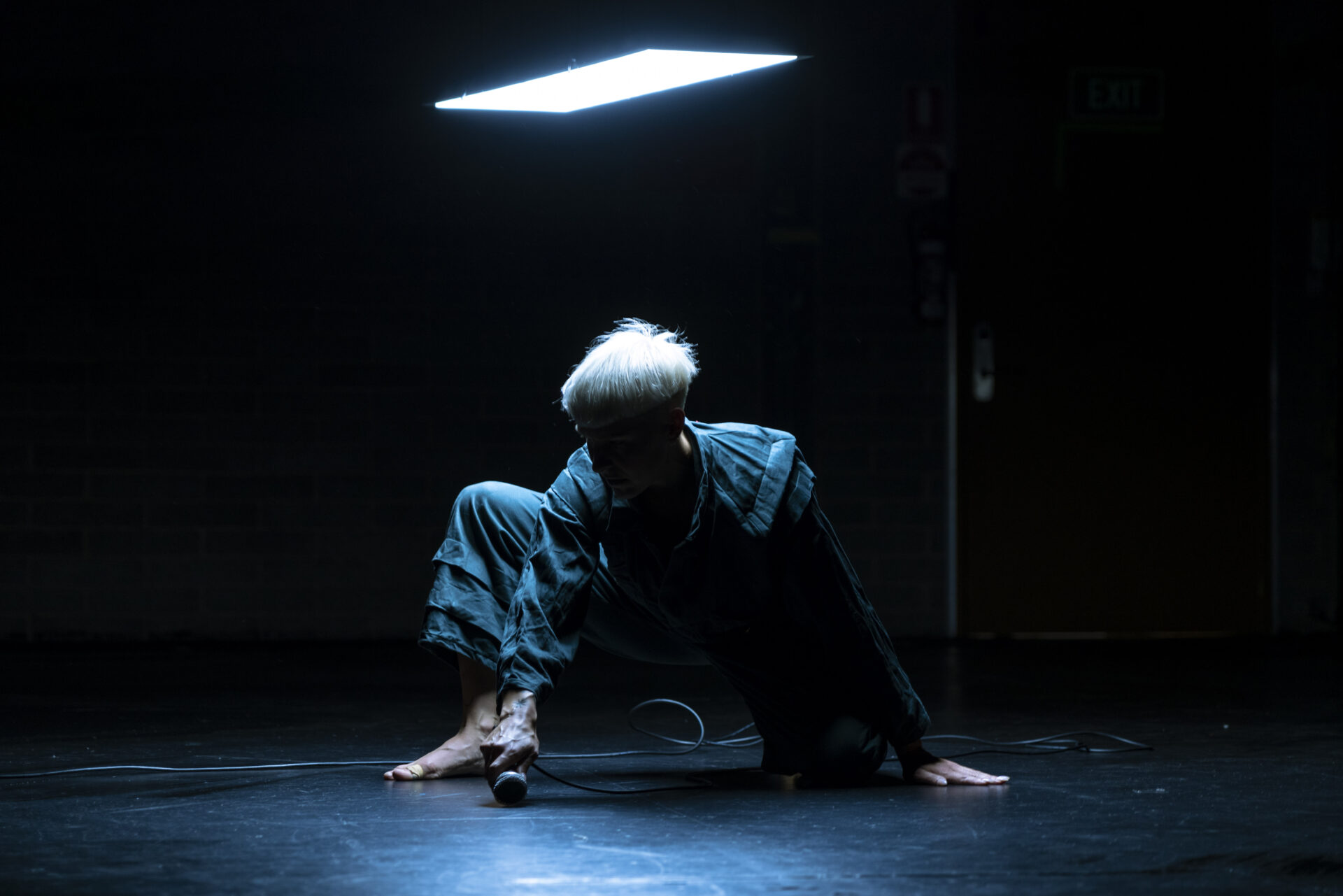 A dancer dressed in a blue boiler suit lunges forward in a dimly lit room during Ashleigh Musk's 'SUB'.