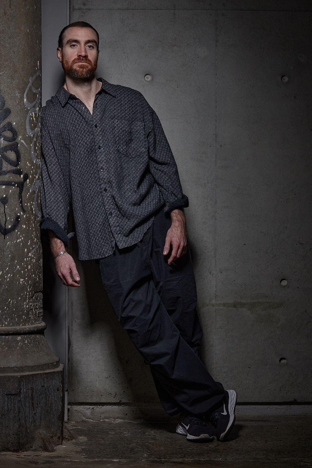 Company Dancer Piran Scott leans against a concrete column dressed in black.